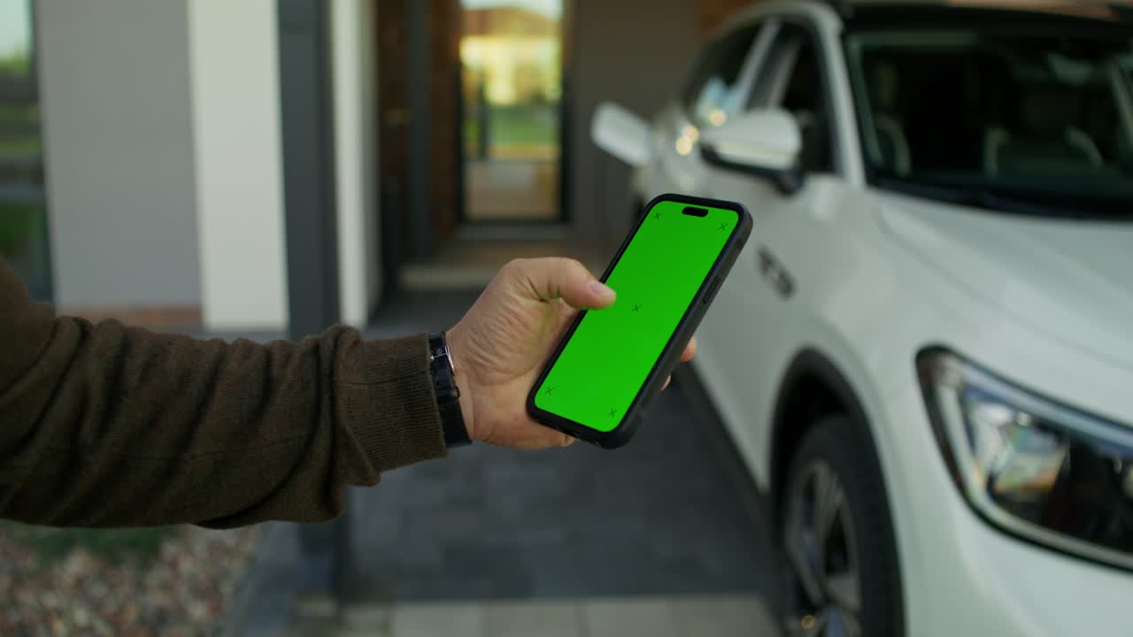 persona sosteniendo un teléfono inteligente con una pantalla verde frente a una casa y un coche