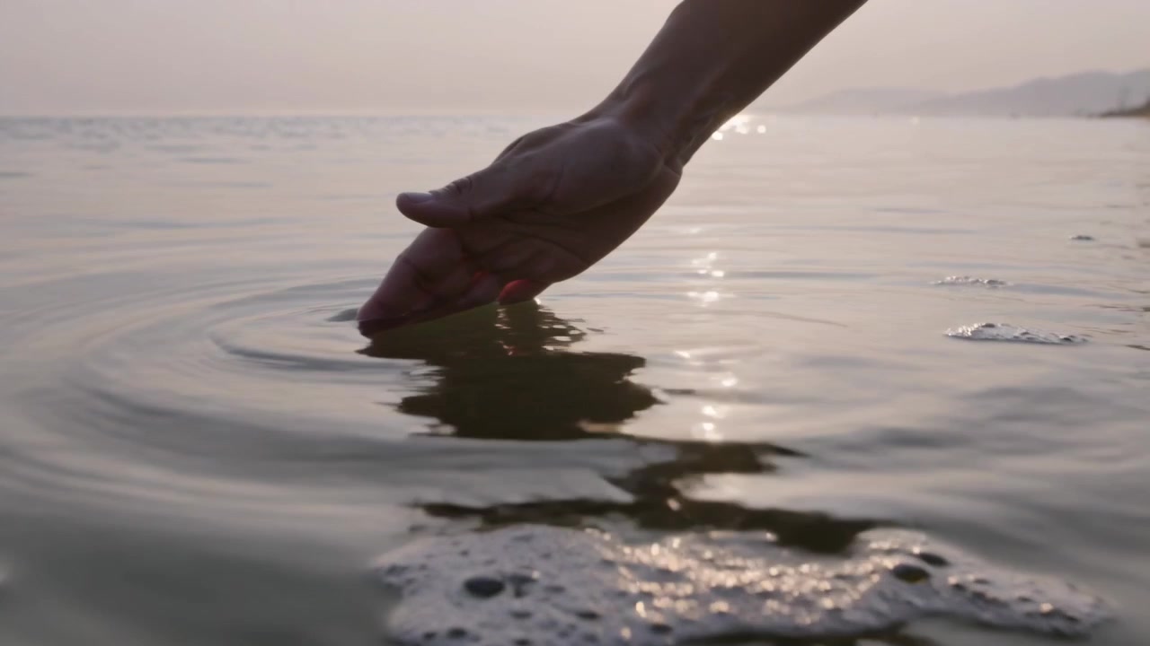 Braccio e mano a maniche corte che entrano dall'alto a destra, sfiorando la superficie del mare e creando increspature, con un riflesso del sole e spazio per il testo.