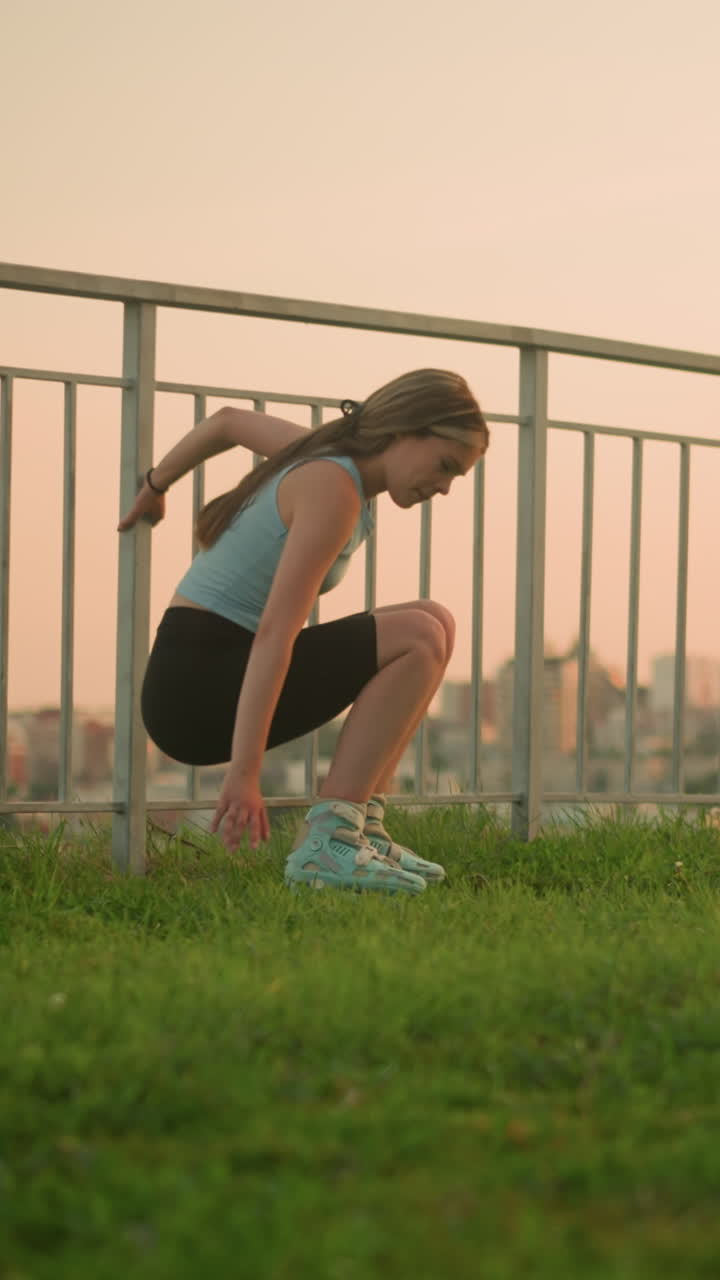 Young lady in roller skates holding iron railing while sitting on grassy ground, with beautiful evening cityscape in blurred background