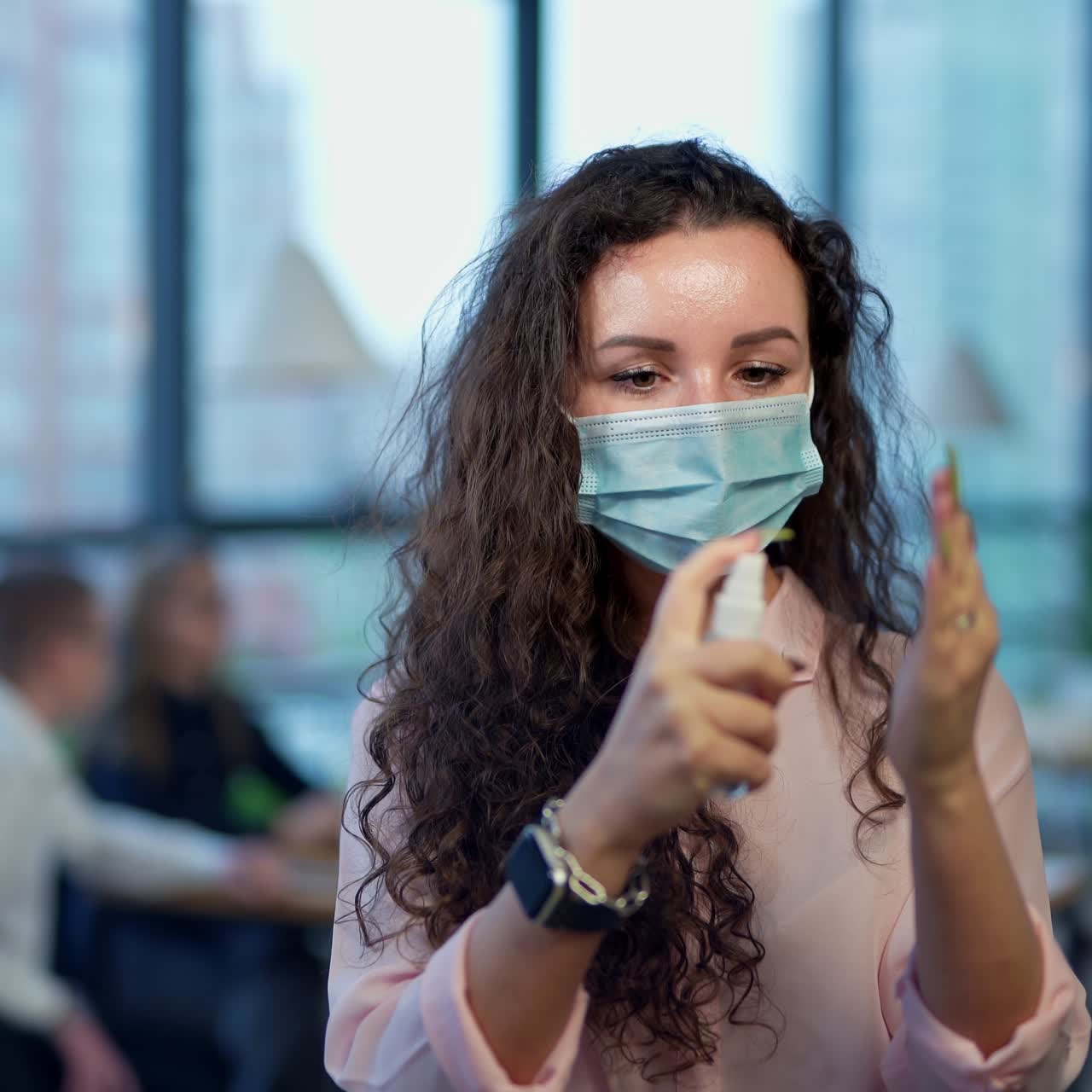 Beautiful young woman putting on a mask and spraying antiseptic. Following anti-pandemic rules at working place. Office staff at the table in blur at the background