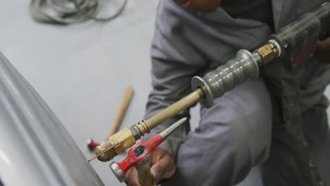 african american male car mechanic holding a screwdriver and using a hammer to repair a car