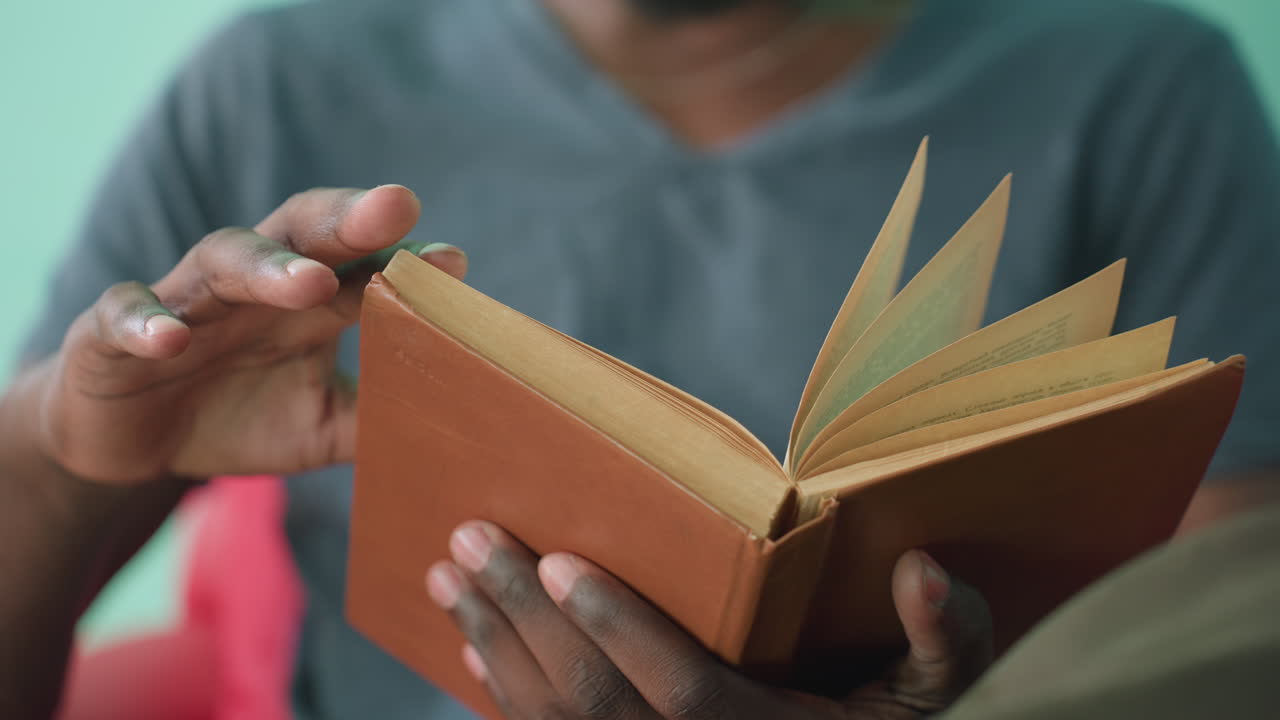 Close up of dark skin student seated on red couch reading worn brown book and flipping to new page, fingers gently turning pages with care, indoors against teal wall in calm quiet atmosphere
