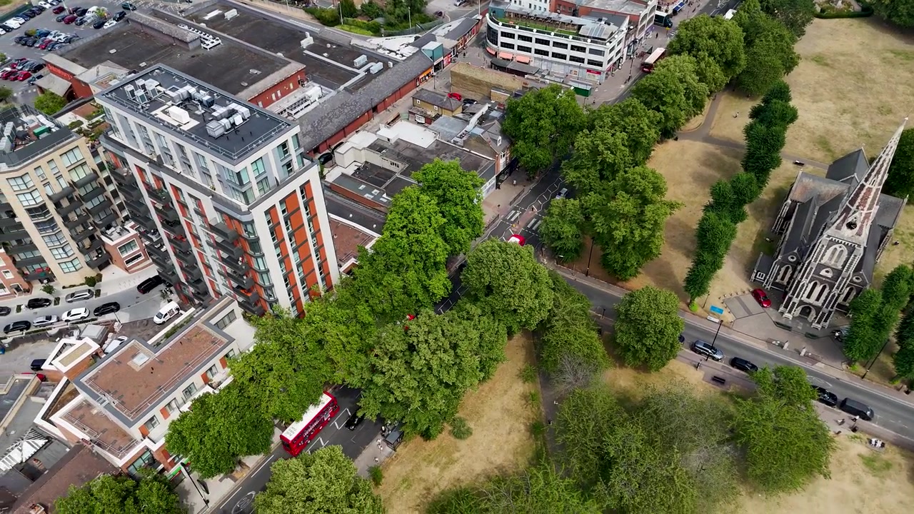Een winkelstraat en een gotische kerk grenzen aan een verdorde gemeenschappelijke weide in een voorstad van West-Londen, met volwassen bomen langs de randen