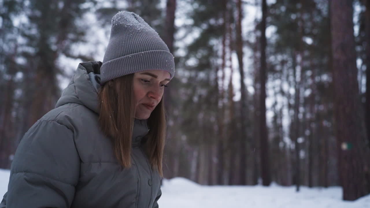 Woman in gray winter coat and knit beanie stands outdoors in snowy forest, appearing focused and contemplative. Snow gently falls around her, capturing serene atmosphere of cold, peaceful winter day