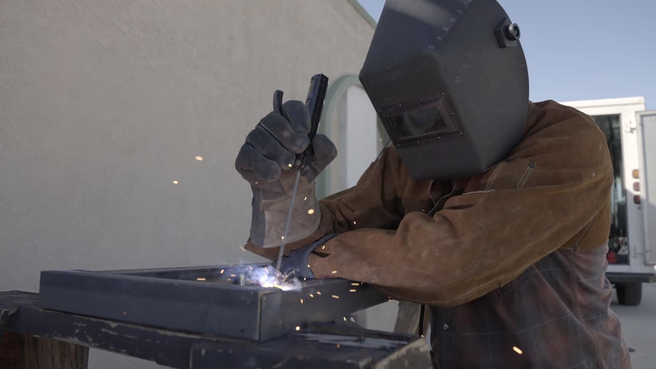 Man wearing welders jacket, gloves, and hood welds angle iron together outside, slow mo