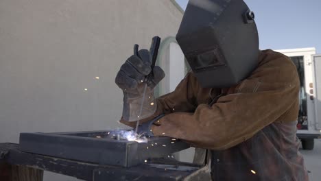 man wearing welders jacket, gloves, and hood welds angle iron together outside, slow mo