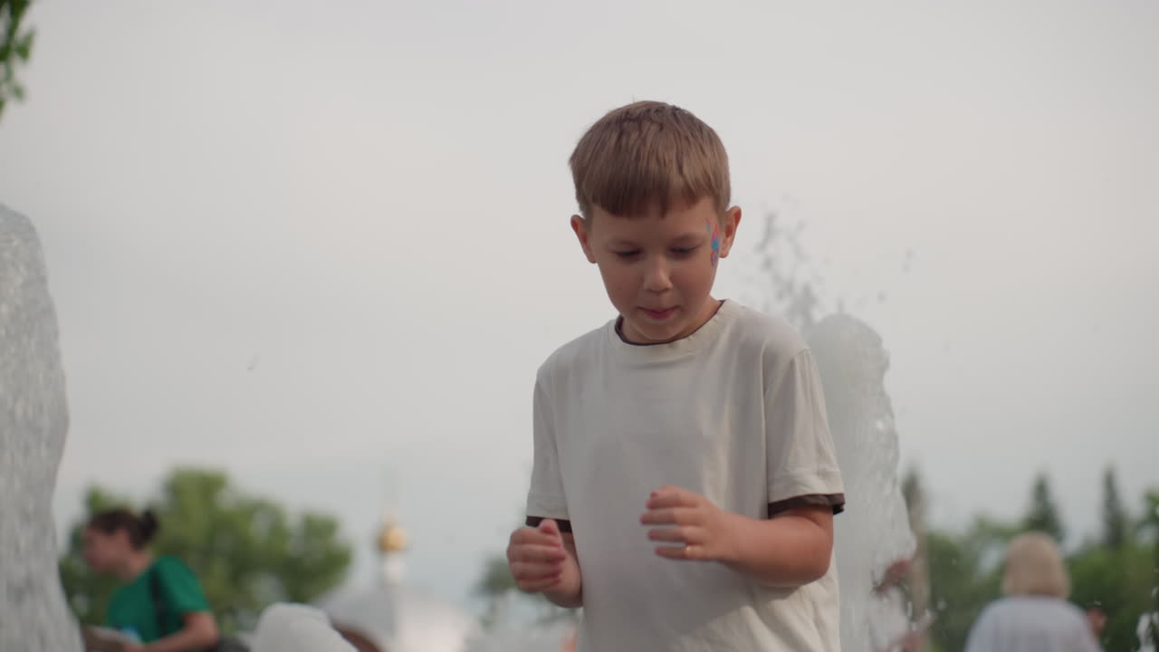 young boy near water fountain turns to playful run from water, smiling with face paint, summer park background with trees and blurred people, splashing droplets catching light