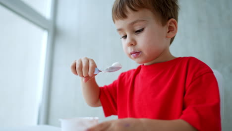 lovely baby boy in red t-shirt eating yogurt from spoon and cup. dairy consuming with appetite. low angle view.