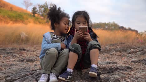 diverse children friend african american and asian girl using smartphone together in nature on mountain with sunlight . group of kid playing internet with mobile smartphone in garden park .