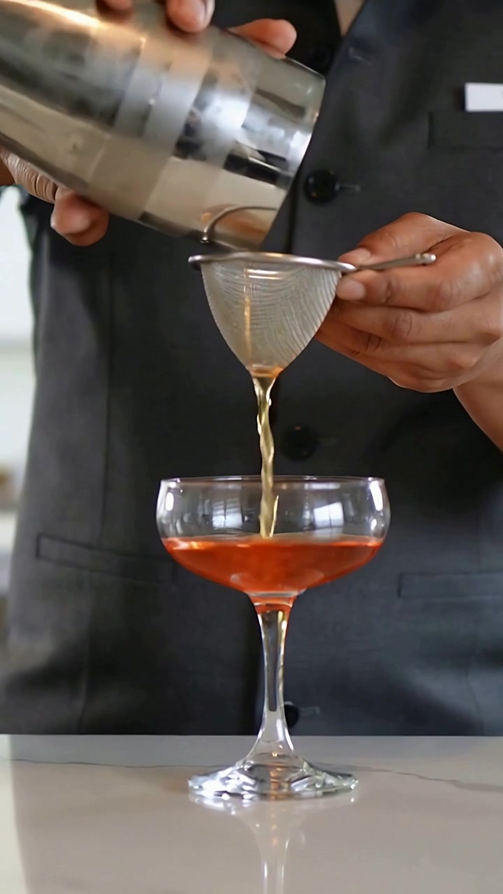Video verticale: Inclinazione dello shaker, barista che filtra la bevanda attraverso un colino in una coppa al bar, etichetta sul gilet