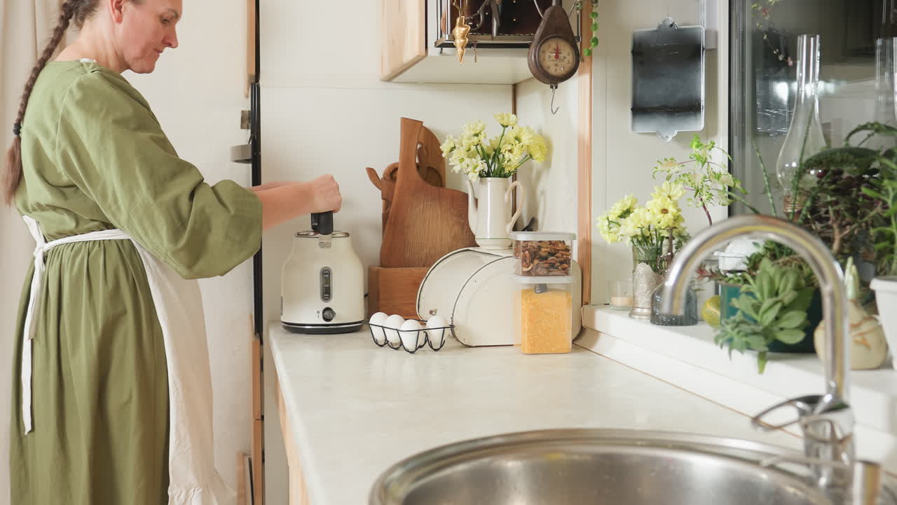 Back view of woman in green dress placing kettle on stove and switching it on as indicator light blinks, surrounded by warm kitchen decor including flowers, eggs, cutting boards, and sink