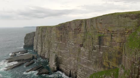 A-slow-panning-shot-revealing-seabirds-flying-over-a-turquoise-green-ocean-in-front-of-a-dramatic-sea-cliff-covered-in-a-seabird-colony-with-waves-crashing-against-its-base