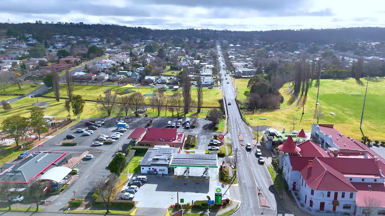 Drone footage tracks above a main road through Armidale suburb, showing moving vehicles, parking lots, and residential areas under bright daylight with scattered clouds
