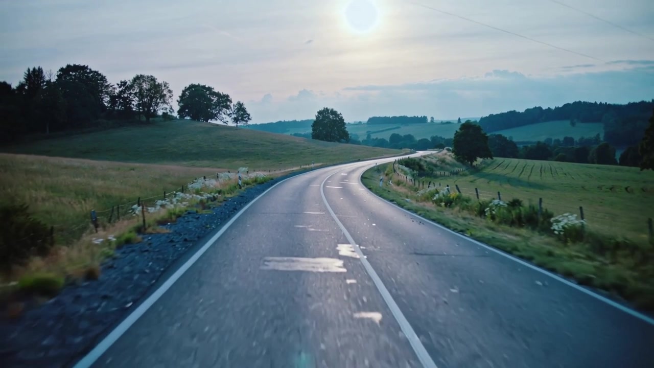 Inquadratura che inizia con la telecamera che si muove lungo una strada rurale verso una curva a destra, mostrando il sole basso e la segnaletica orizzontale.