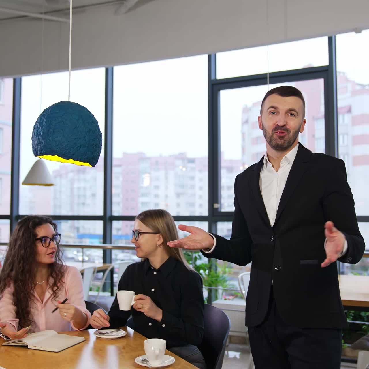 Businessman giving a talk to the camera and gesturing with his hands. Team of workmates sit next at the table having a conversation