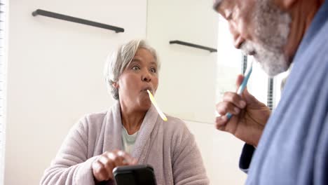 pareja mixta racial feliz cepillándose los dientes, usando el teléfono inteligente en el baño, sin alteración, en cámara lenta