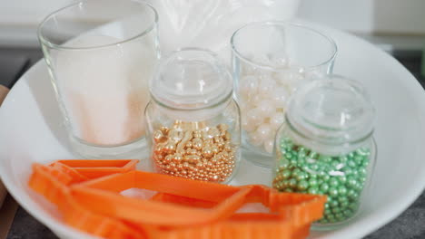 close-up of colorful decorative beads in glass jars, sugar in glass cup, and orange baking tools placed in white bowl on kitchen countertop, sparkling edible pearls and sugar crystals