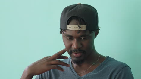 close up of young man wearing black cap backward and casual shirt, touching face gently with thoughtful expression as he studies, nodding subtly in focus against soft teal background