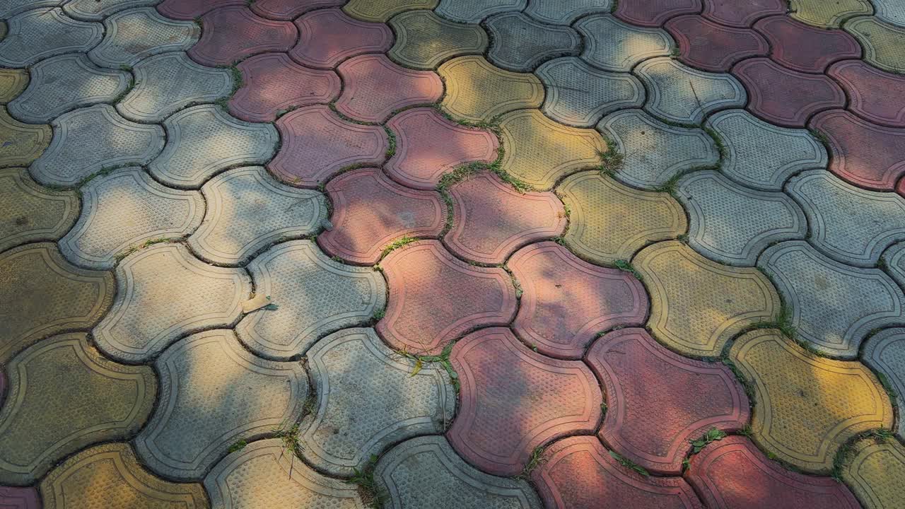 A slow tracking view over patterned interlocking tiles in red, yellow, and gray, softly lit by shifting tree shadows that create gentle contrasts on the ground