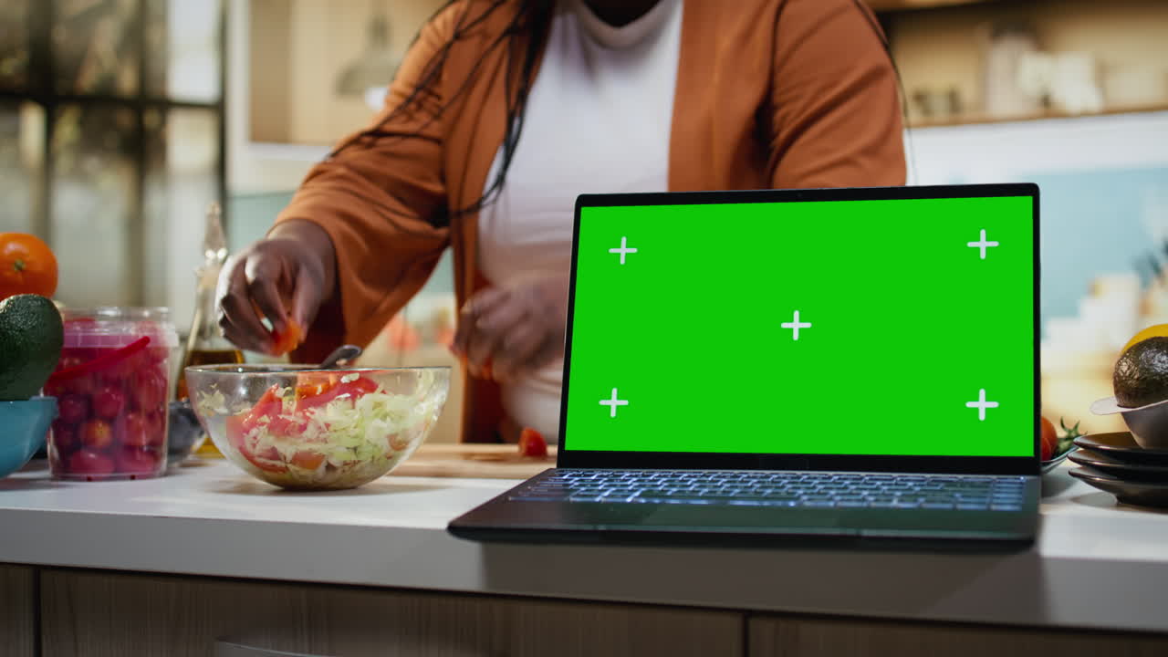 Schermo verde su laptop accanto a una donna nera che registra la preparazione dei pasti per una ricetta.