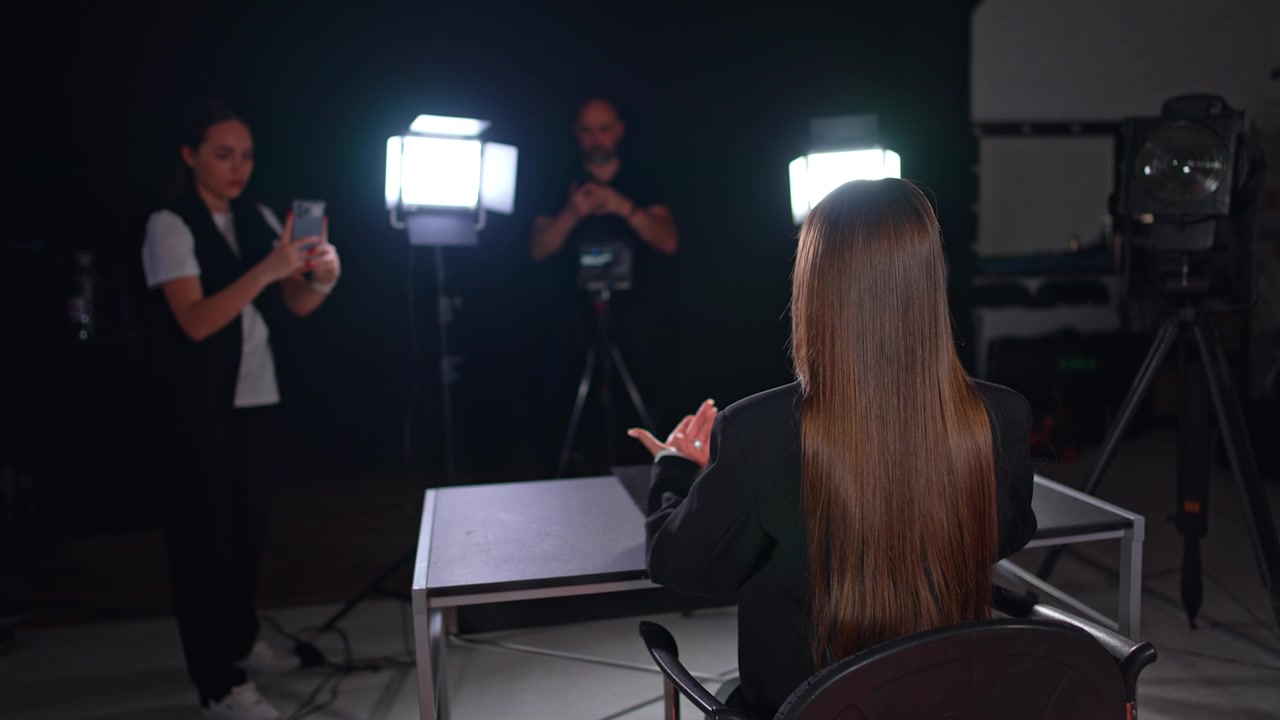 Vista posteriore di una blogger dai lunghi capelli castani che indossa una giacca nera seduta alla scrivania. Donna e uomo sullo sfondo che girano video con diversi dispositivi