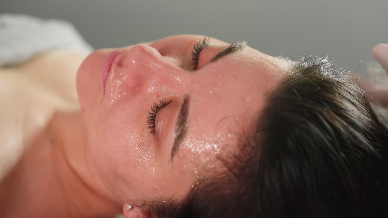 Close Up Spa Visitor Lying With Eyes Closed On Bed While Beauty Technician In Gloves Dips Brush Into Clear Gel And Applies Nourishing Face Mask In Gentle Strokes For Hydrated, Radiant Skin Treatment