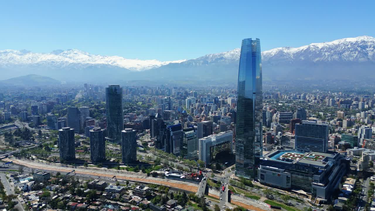 Drone aerial ascending past the glass Costanera tower, revealing downtown skyline and snowcapped Andes under clear daytime light. Santiago, Chile