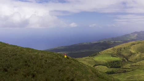 Asian-Malaysian-Chinese-tourist-woman-making-video-with-phone-on-a-path-on-the-edge-of-the-vulcanic-lush-green-mountain,-on-Pico-da-Esperança,-drone-dolly-in-São-Jorge-island,-the-Azores,-Portugal