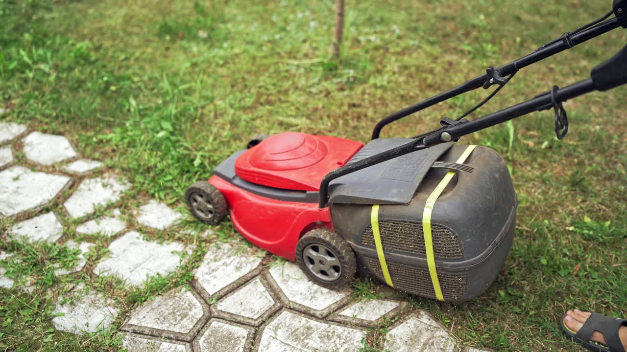 A lawn mower is cutting green grass. Young boy with a lawn mower is working in the backyard