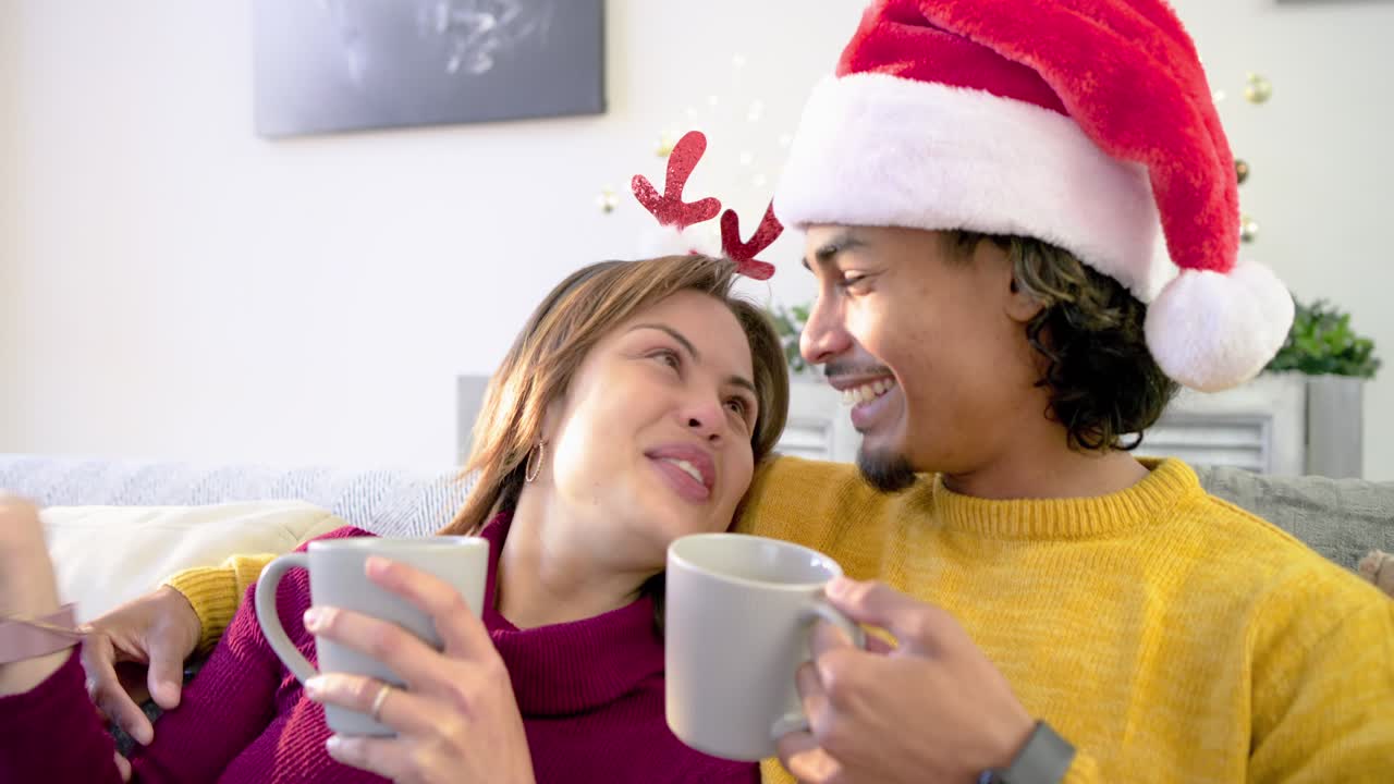 Garland inviting Diverse couple settling onto sofa in living room, toasting grey mugs and laughing