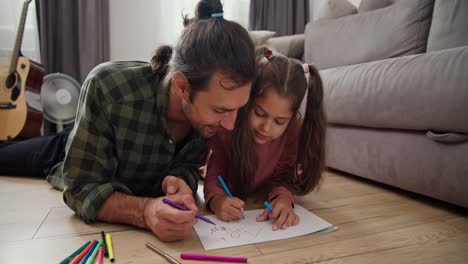 a lonely father, a brunette man in a green checkered shirt lies on the floor with his little daughter in a pink dress and draws on a sheet of paper using multi-colored felt-tip pens near a gray sofa in a modern apartment