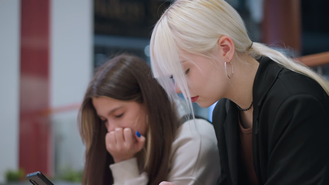 Young ladies seated indoors focused on mobile gadgets with serious expression showing youth lifestyle digital culture technology dependence modern communication