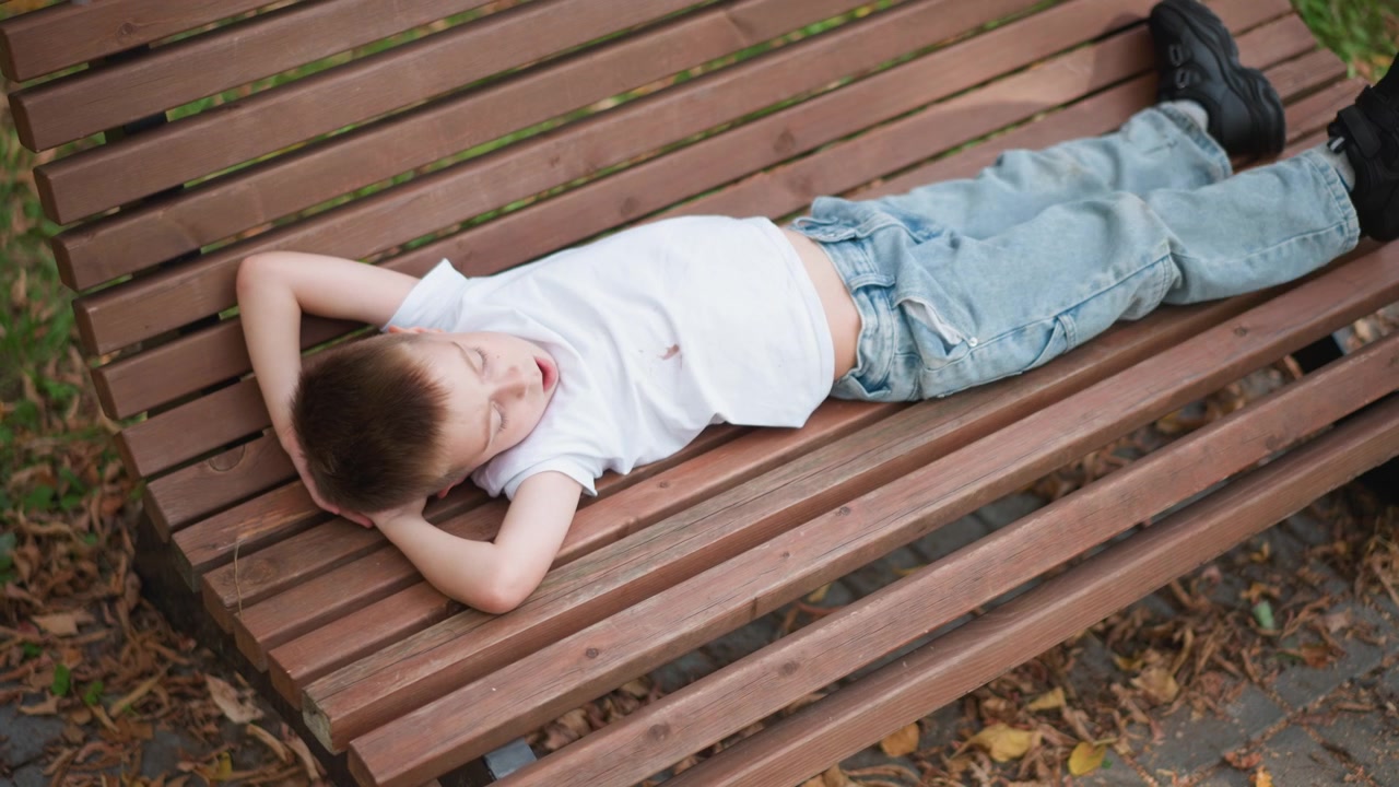 Young boy lies on wooden bench in outdoor park setting appearing to rest or sleep with his arms behind his head. Boy wears white t shirt and light blue jeans background shows green grass