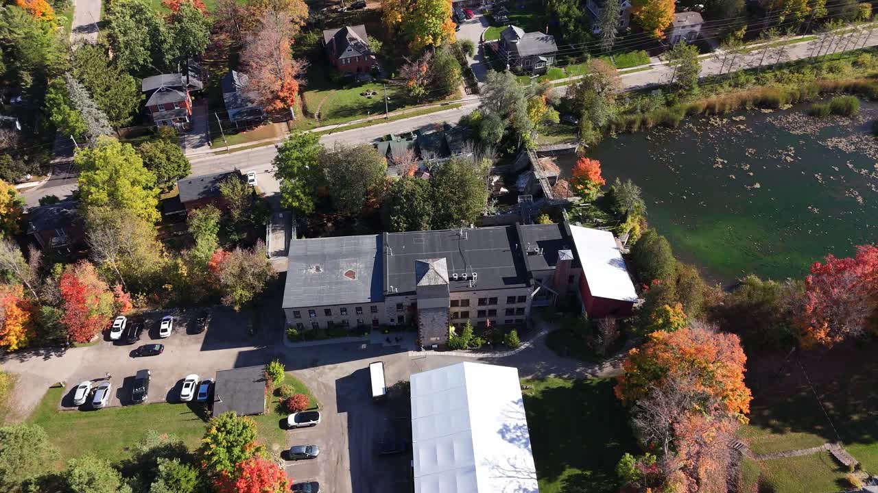 A detailed aerial view of the historic Alton Mill Arts Centre in Ontario, Canada. The shot showcases the stone building and its surroundings in peak autumn colour
