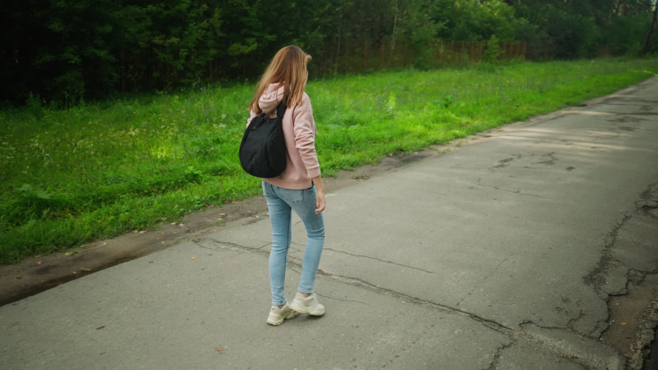 Girl with loose hair walking thoughtfully along forest path with backpack, surrounded by fresh green grass and forest trees, cracked pavement beneath her feet