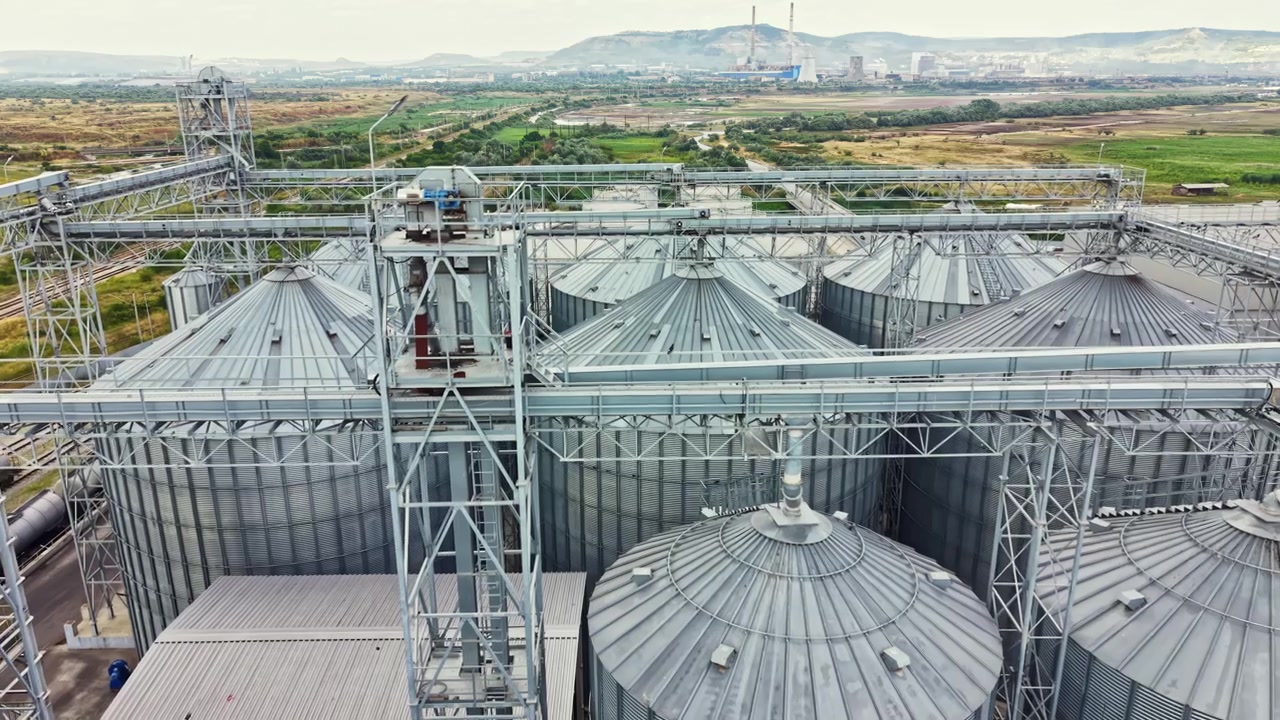Farm storage units and silos located in an agricultural area show machinery and activity in the background on a cloudy day