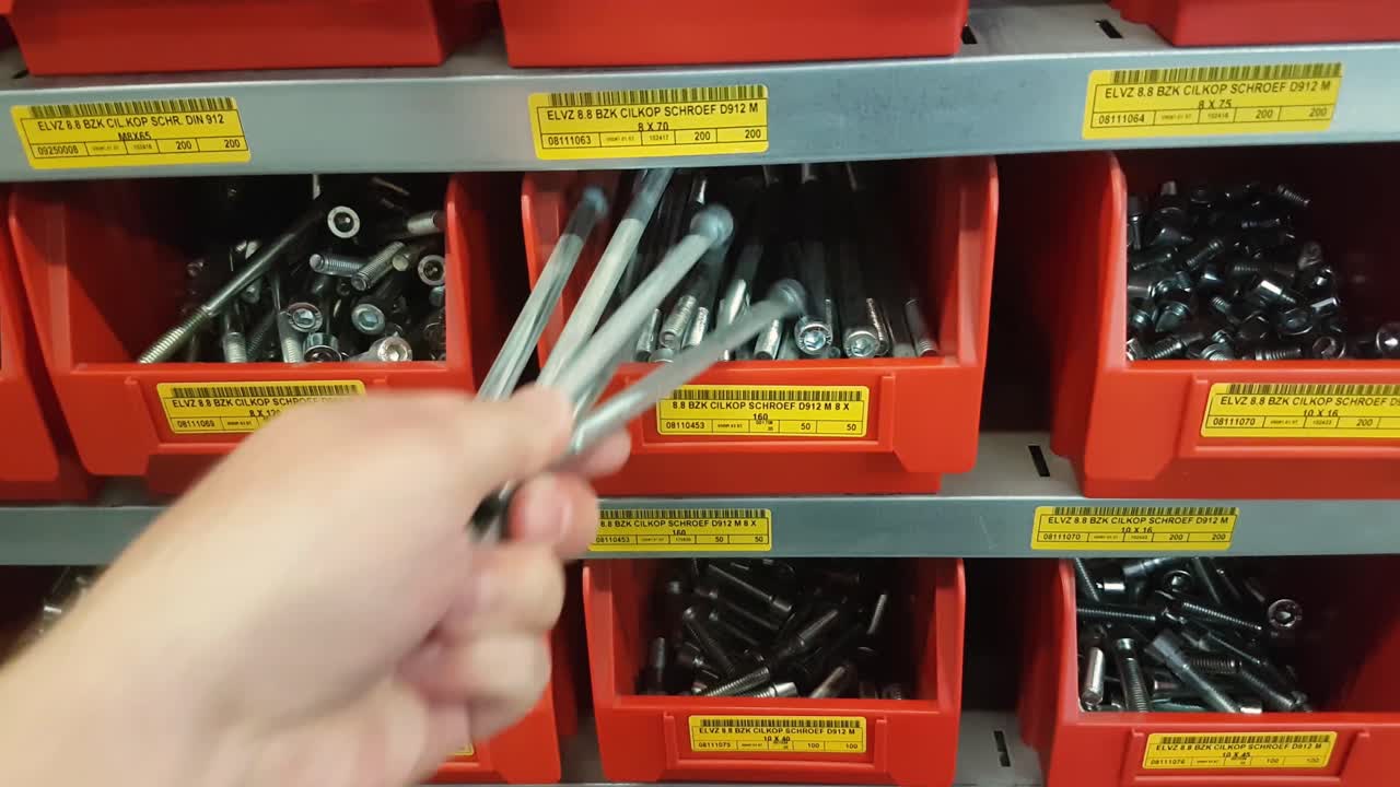 A human hand takes out five large bolts from a red storage bin in a steel industrial rack. CLOSE UP