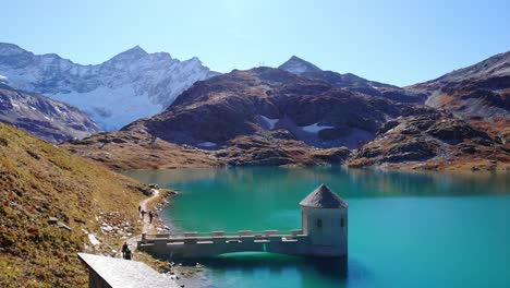 Aerial-View-Of-Hikers-Walking-On-The-Trail-With-Intake-Tower-At-Weisssee-Reservoir-In-Austria