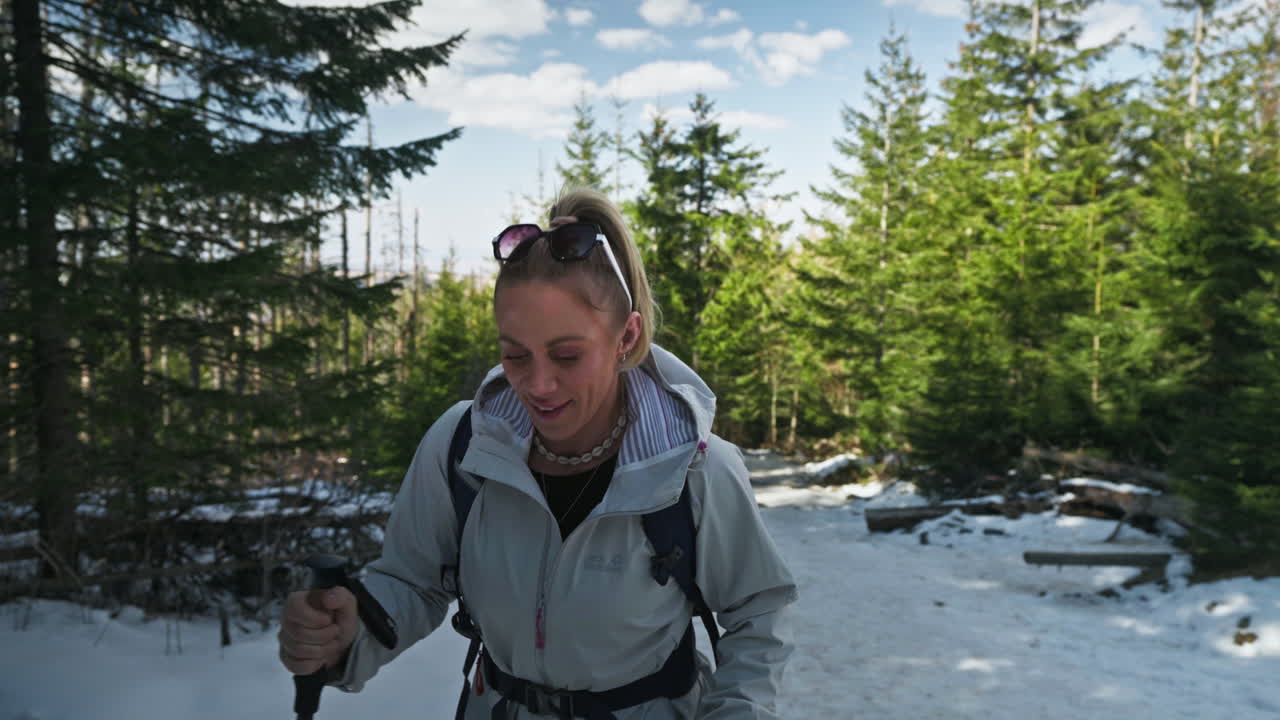 A woman walks uphill using hiking poles on snow covered forest trail with clear blue sky above, slow motion frontal view, medium