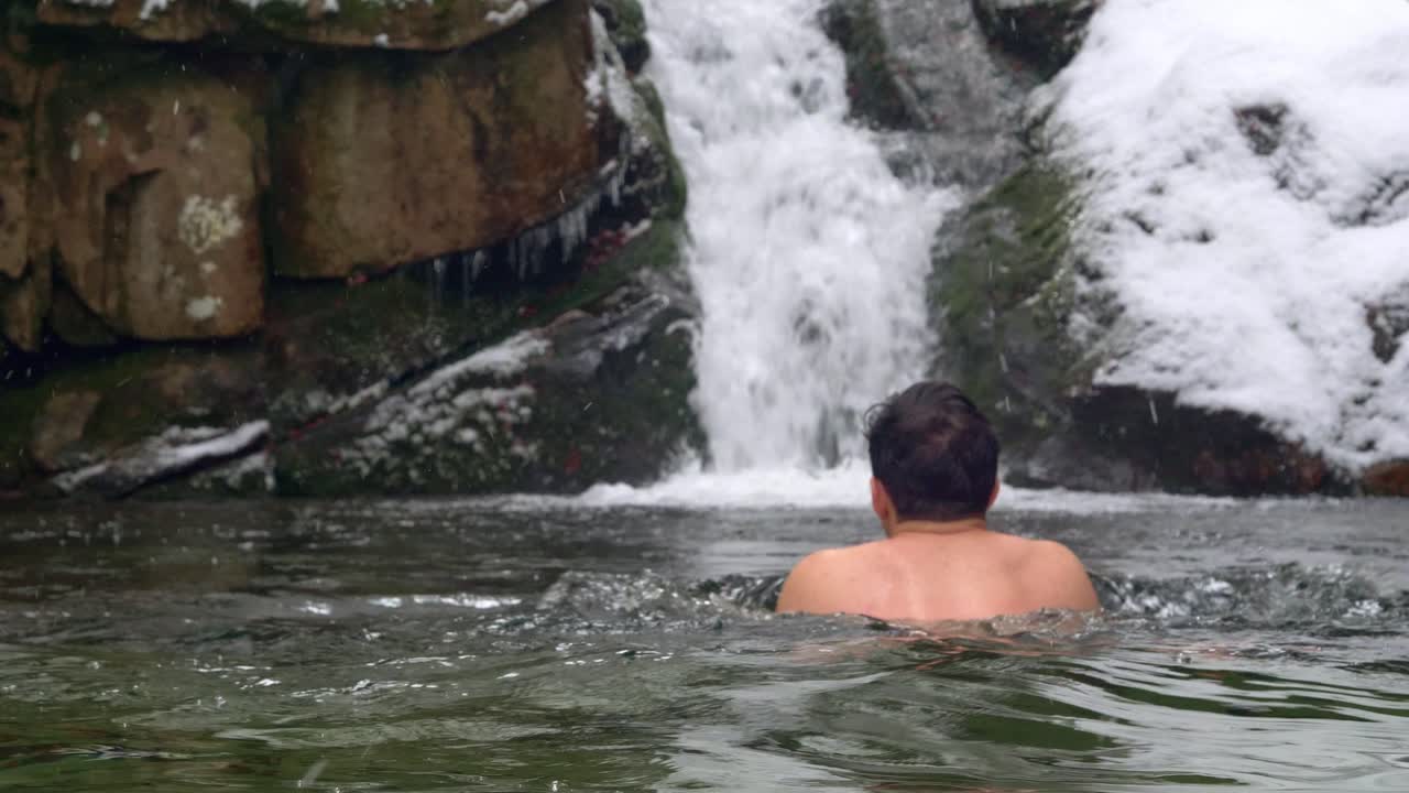 흐린 겨울날 눈 덮인 산의 차가운 강에서 폭포를 향해 수영하는 용감한 남자 얼음 - 정적 샷, 느린 동작