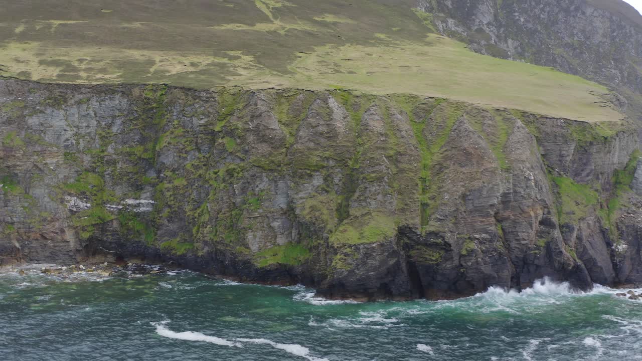 Mossy sheer sea cliff with bright green grass and strong waves crashing on basalt, achill island ireland