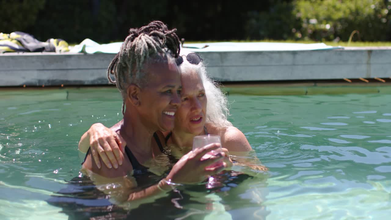 Femme âgée parlant et souriant, sa compagne se relaxant dans une piscine, tenant des boissons roses.