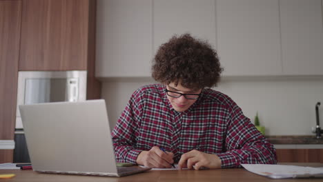 curly haired male student attractive young boy in glasses is studying at home using laptop typing writing in notebook. college student using laptop computer watching distance online learning seminar