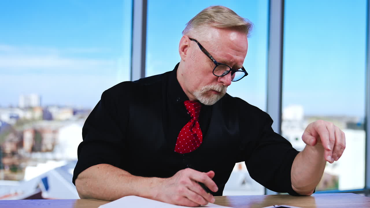 Busy serious man wearing black clothes and red tie works at desk. Man uses I-pad and takes notes into his paper notebook.