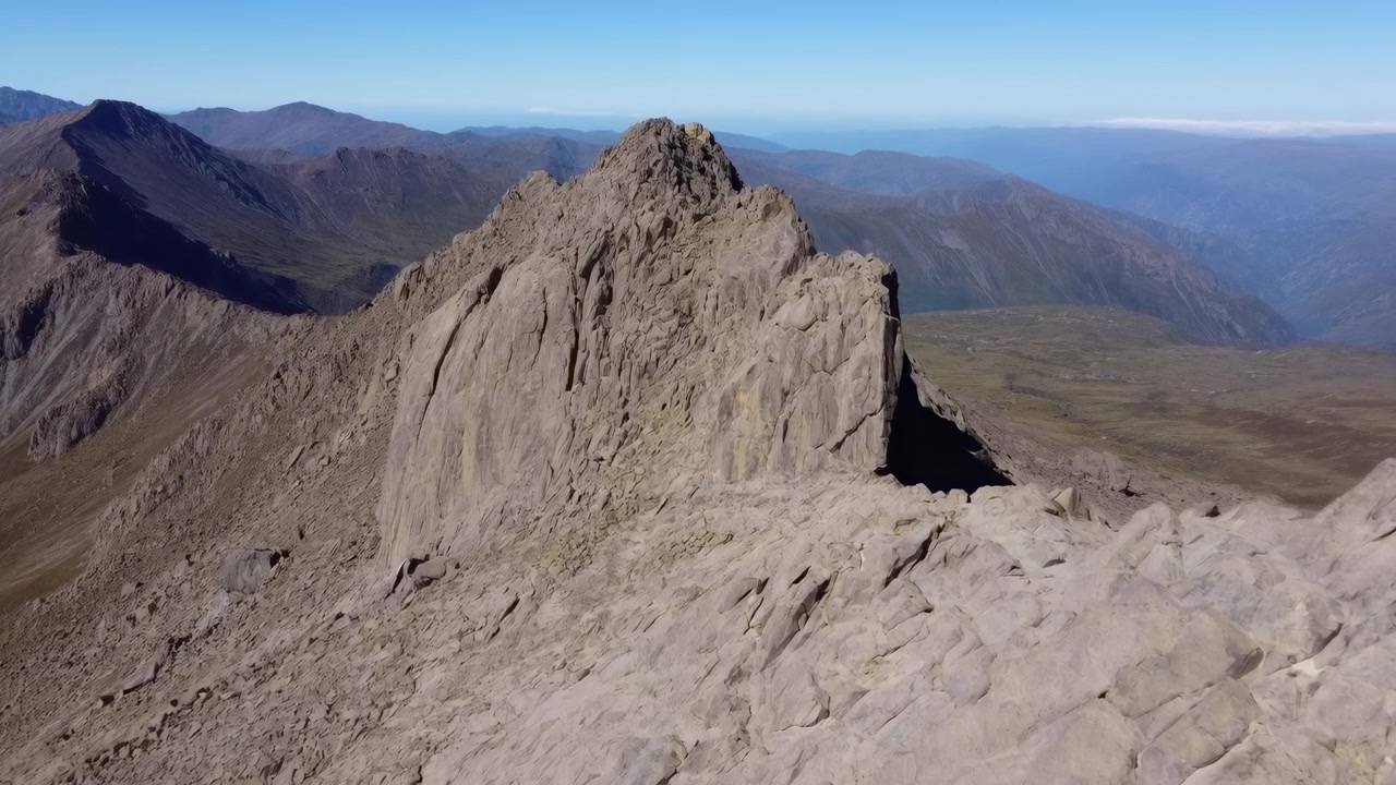 Vista aerea con drone: cima di montagna rocciosa e cresta di pietra sotto un cielo azzurro limpido, valle distante e colline ondulate.