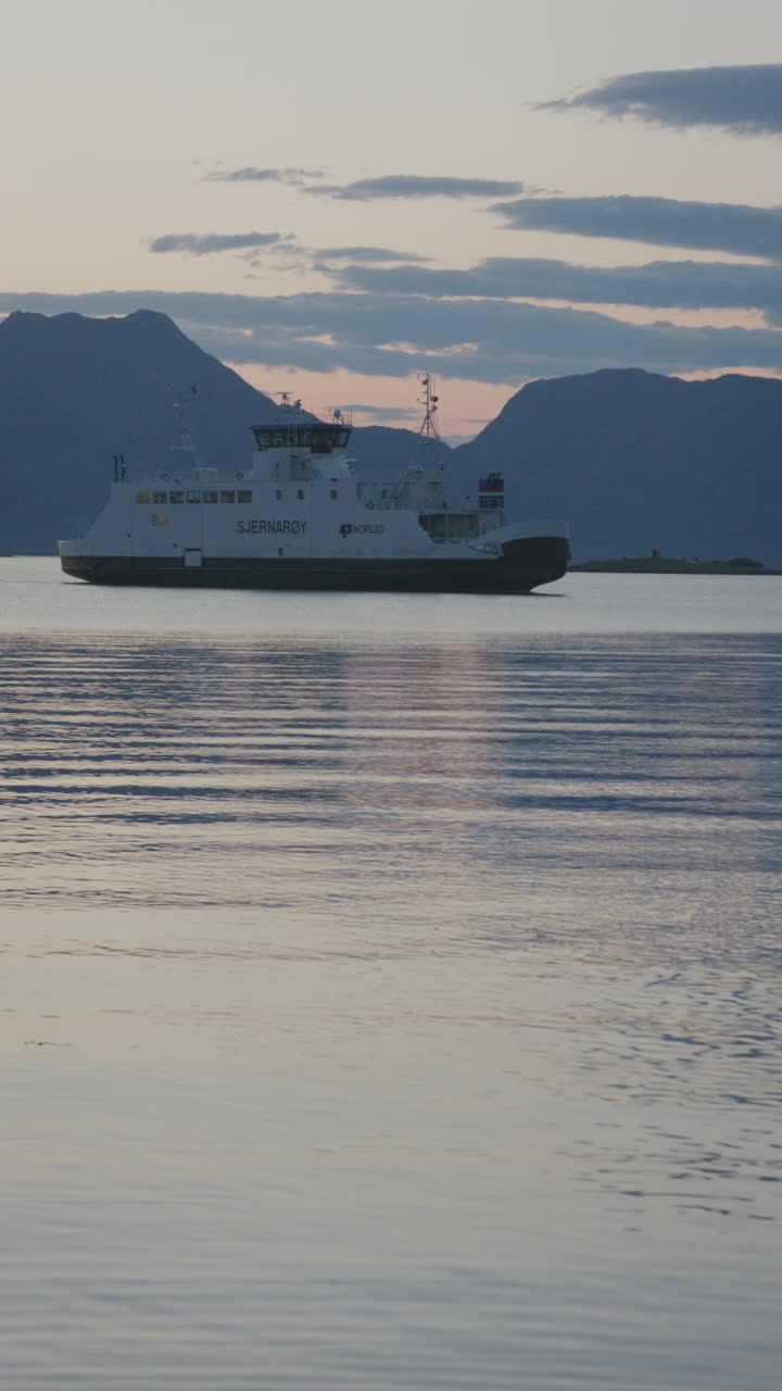 A vertical shot of a ferry sailing across still waters at dusk, framed by mountains.