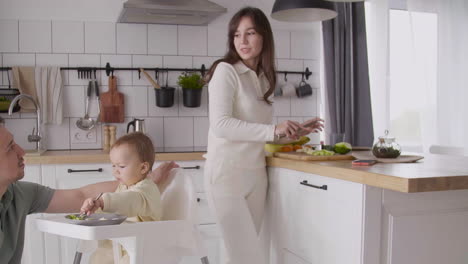 father feeding his happy baby girl sitting in her high chair in the kitchen while his wife cutting fruit and talking with him