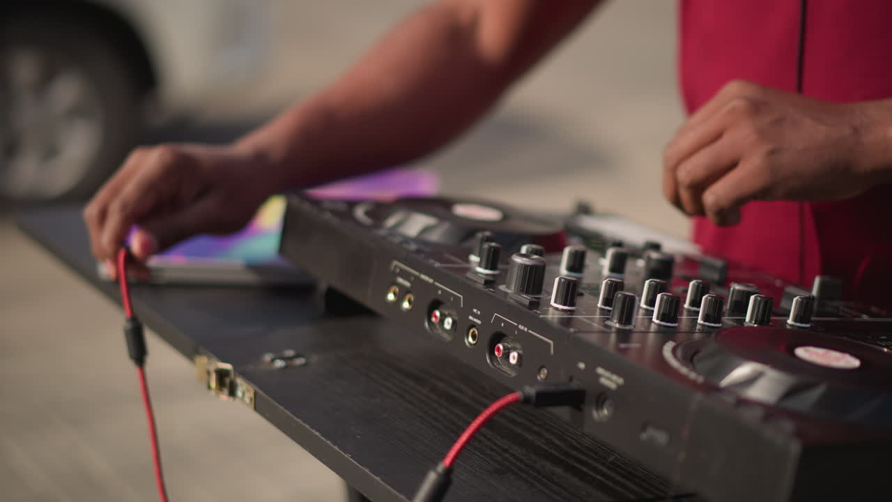street DJ mixing on portable controller, closeup hands adjusting knobs and fader, connecting red cable to mixer, vinyl sleeve beside folding table, outdoor daytime sidewalk scene, casual performance