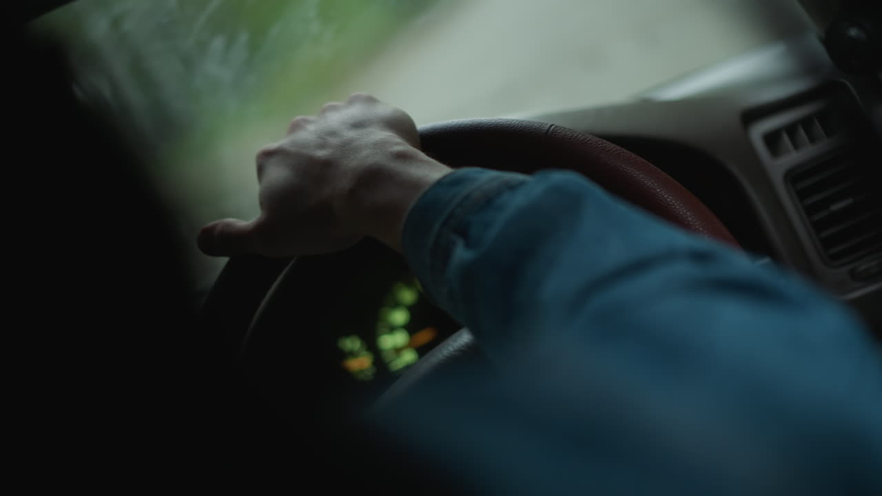 Hvit mann som kjører bil og sjekker instrumentene på dashbordet under et svakt, grønt indikatorlys, rød varsellampe, hender som justerer grepet, våt frontrute, forsiktig navigering gjennom bygater