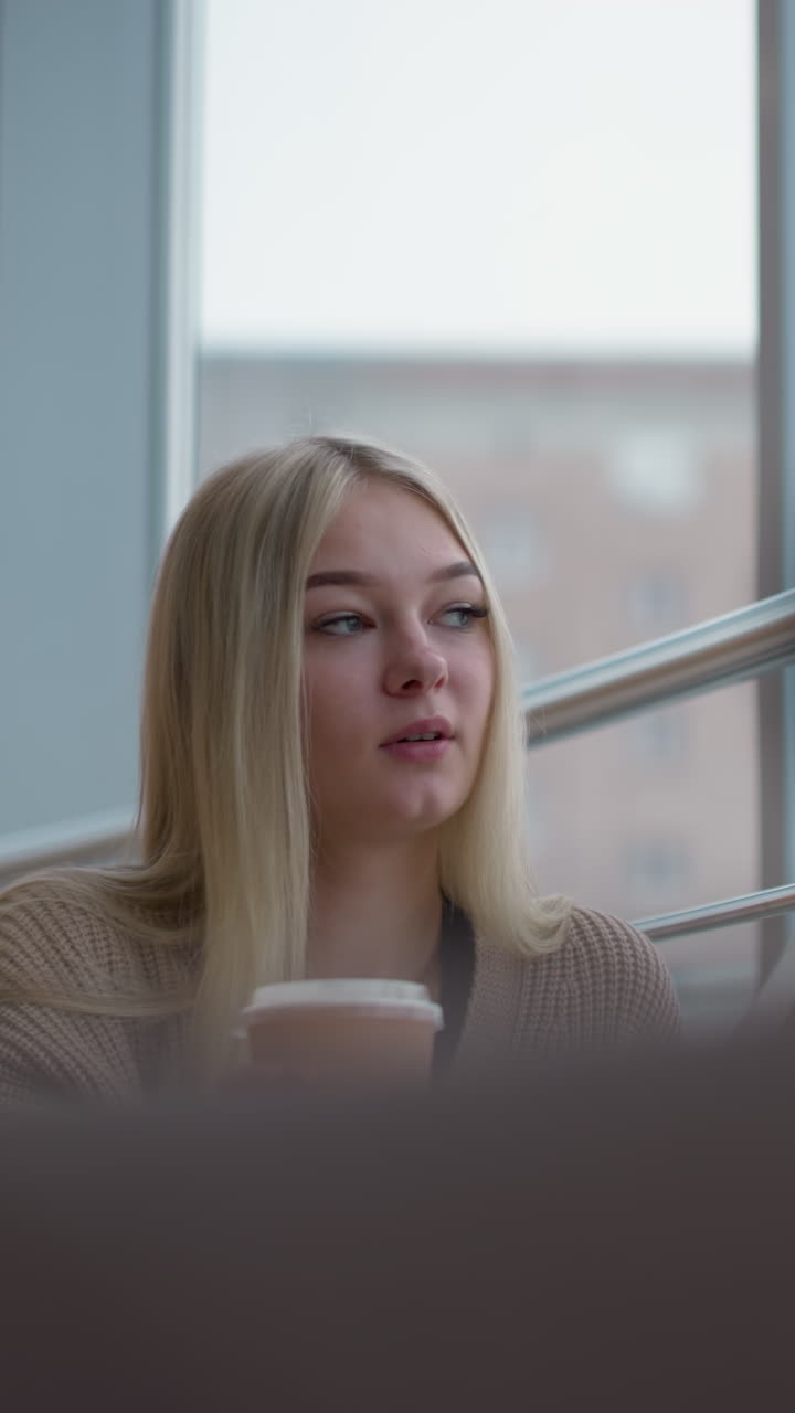 Ung dame som nipper til te fra koppen sin, mister den på bordet mens hun ser ut gjennom gjennomsiktig glass i et kjøpesenter, med mykt lys som filtreres gjennom glasset.